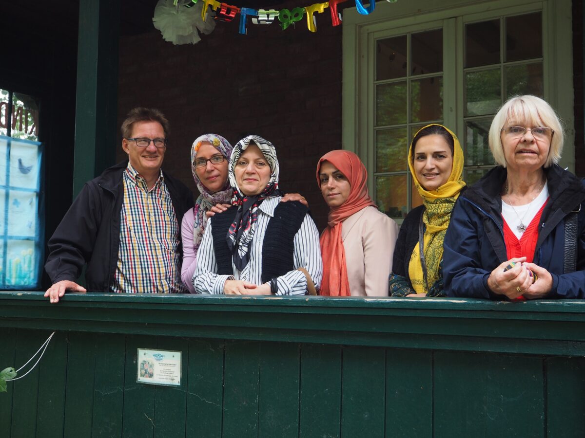 Sechs Personen stehen auf einer Veranda mit grüner Holzbrüstung, einige tragen Kopftücher, darüber bunte Girlande mit Buchstaben