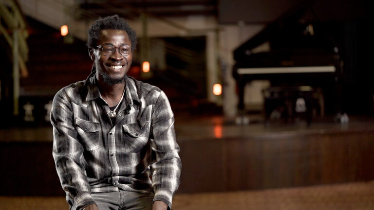 Ein dunkelhäutiger Mann mit kariertem Hemd lächelt und schaut aus dem Foto.