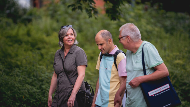 Eine Frau und zwei Männer während einer Unterwegsgesprächsrunde von Sprachbrücke-Hamburg e.V. spazieren. Im Hintergrund ist eine Hecke oder Bäume zu sehen.