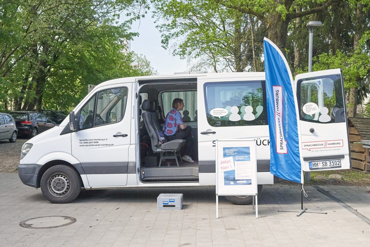 Weißer Sprinter seitlich zu sehen mit offener Schiebetür. Innen eine Gesprächsleiterin von Sprachbrücke-Hamburg e. V. sitzend. Davor ein Plakataufsteller und ein Werbesegel
