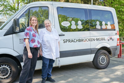 Eine jüngere und eine ältere Frau stehen vor einem weißen Kleintransporter, auf dem "Sprachbrücken-Mobil" steht.
