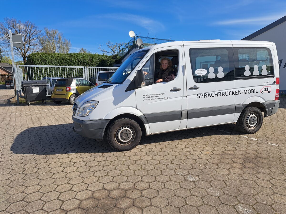 Ein weißer Kleintransporter seitlich steht auf einem großen Platz. Am Fahrersitz sitz eine Frau und lächelt in die Kamera