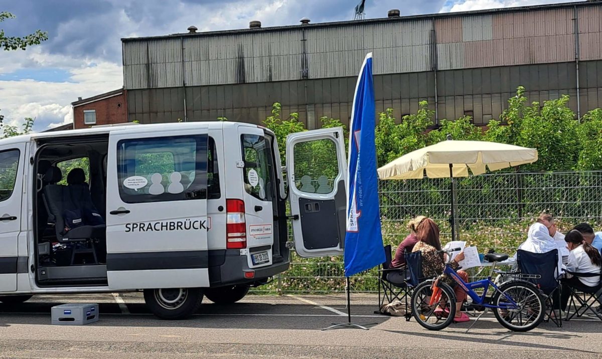 Ein weißer Kleintransporter steht mit offener Seiten- und HIntertür auf einem großen Platz. Daneben sitzen Menschen unter einem Sonnenschirm im Stuhlkreis und unterhalten sich.