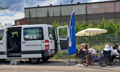 Ein weißer Kleintransporter steht mit offener Seiten- und HIntertür auf einem großen Platz. Daneben sitzen Menschen unter einem Sonnenschirm im Stuhlkreis und unterhalten sich.