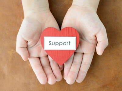 Ein Holzherz mit der Aufschrift Support liegt in 2 Handflächen