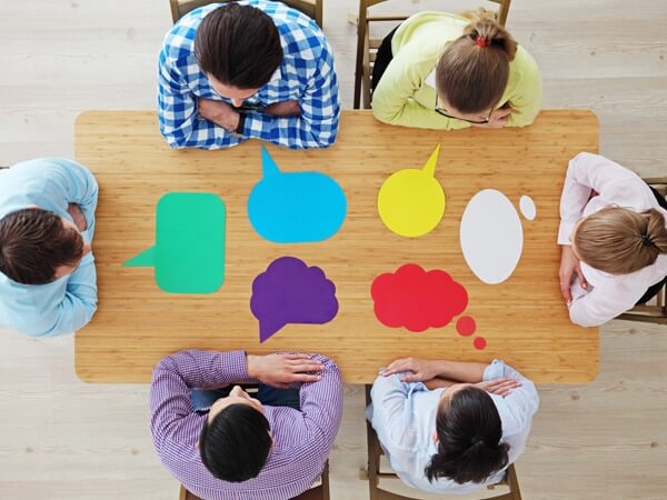 Menschen sitzen an einem Tisch, vor ihnen bunte Sprechblasen