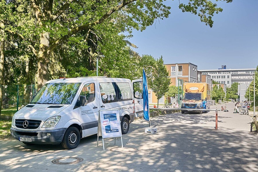 Weißer Sprinter seitlich zu sehen mit offener Schiebetür. Innen eine Gesprächsleiterin von Sprachbrücke-Hamburg e. V. sitzend. Davor ein Plakataufsteller und ein Werbesegel. Im Hintergrund ein Gebäude