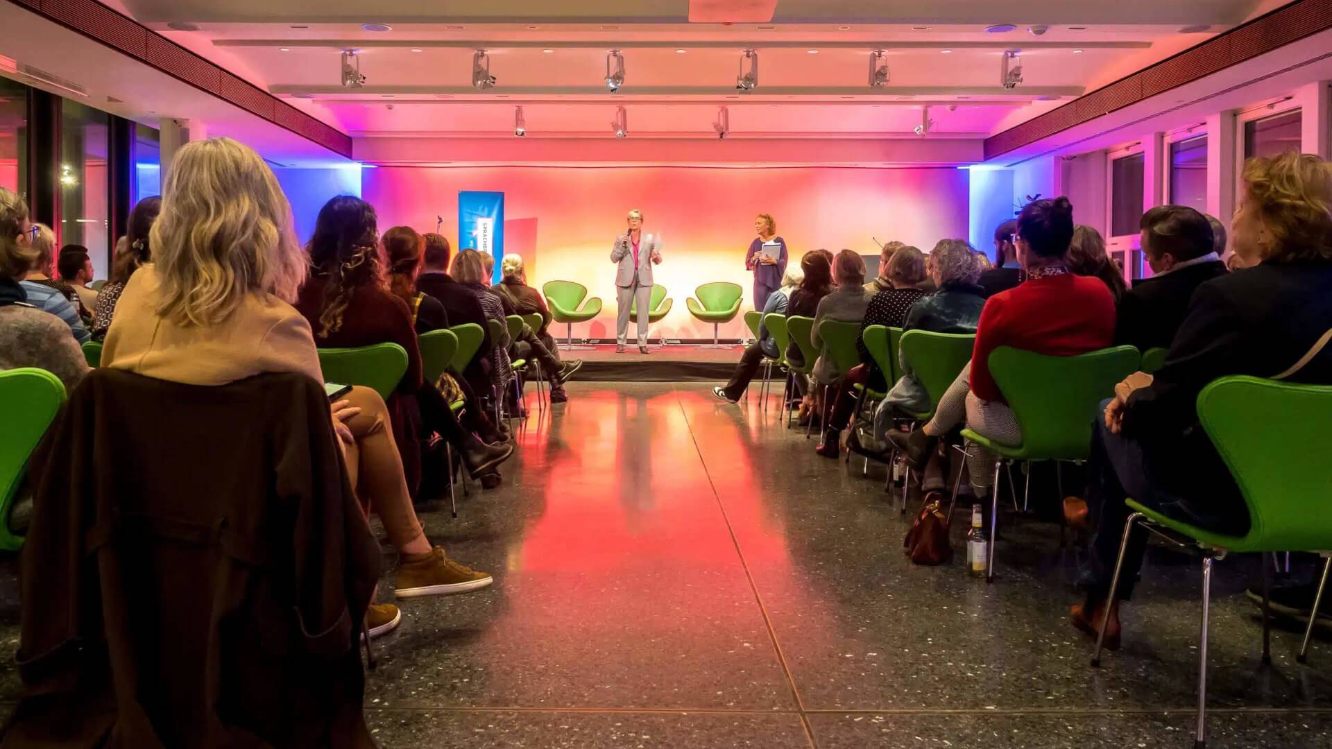 Veranstaltungssaal, in dem Menschen vor einer Bühne sitzen. Auf der Bühne zwei Rednerinnen.